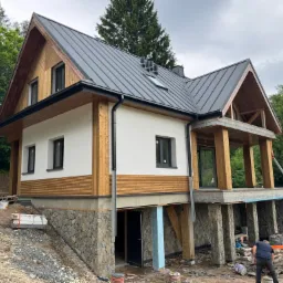 Dwukondygnacyjny dom z dachem z blachy na kamiennej podmurówce, otoczony zielenią. Widoczne elementy drewniane wykończenia i prace wykończeniowe na zewnątrz.