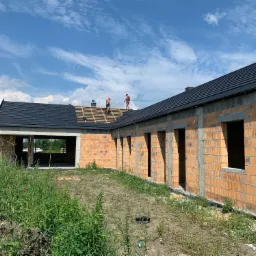 Budowa domu jednorodzinnego w stanie surowym otwartym: widoczne mury z pustaków ceramicznych, przygotowane otwory okienne i dach z czarnej dachówki, na którym pracują dwaj dekarze w czerwonych...