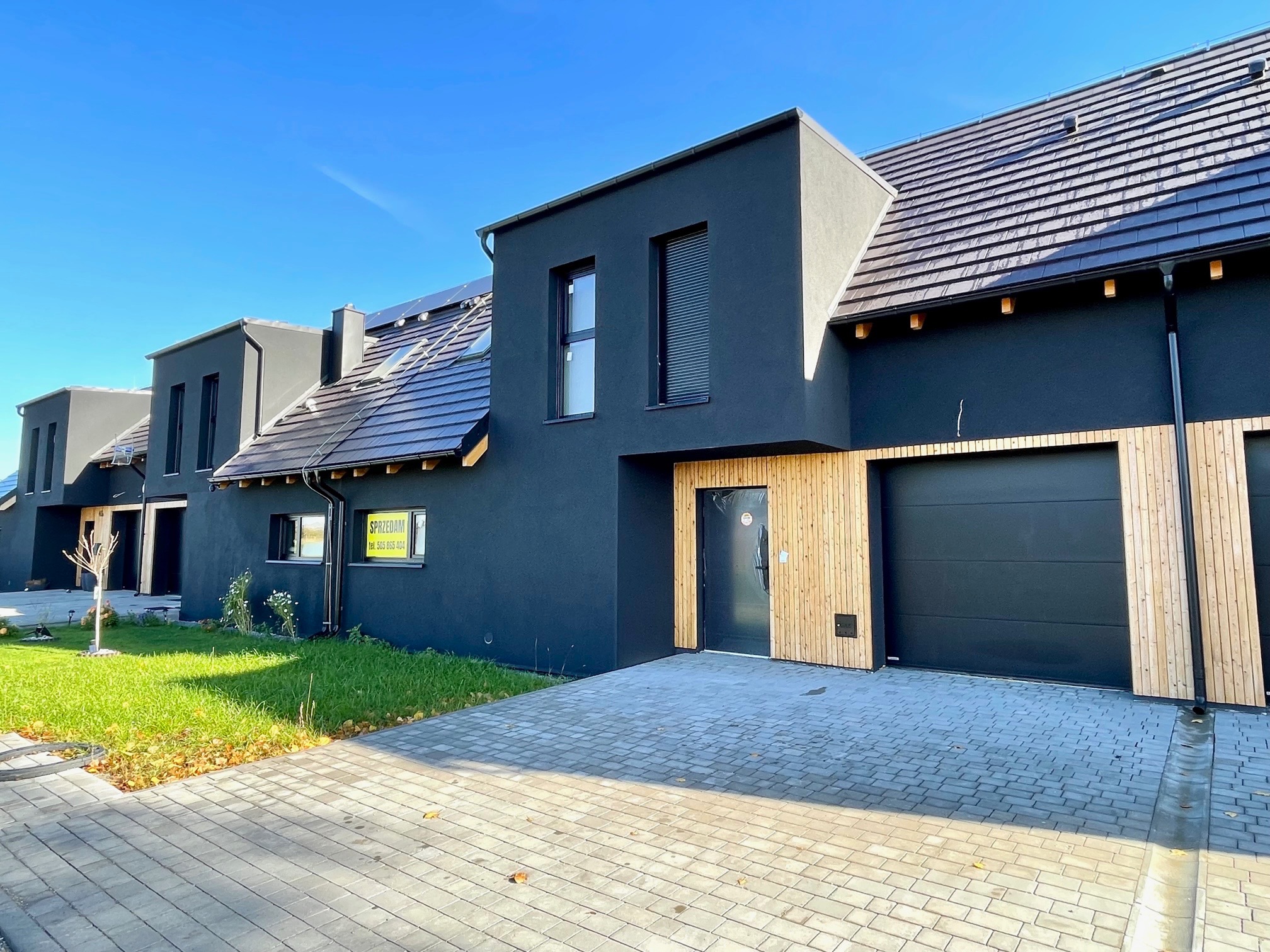 Szeregówka nowoczesnych, czarnych domów z drewnianymi akcentami elewacji i kostką brukową na podjeździe, w słoneczny dzień.