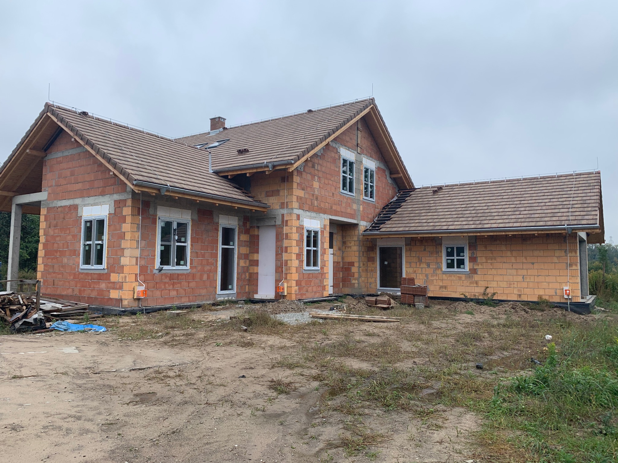 Surowy stan domu z cegły ceramicznej z częściowo położonym brązowym dachem, widoczne białe ramy okienne i drzwi, teren wokół domu niezagospodarowany, pochmurne niebo.