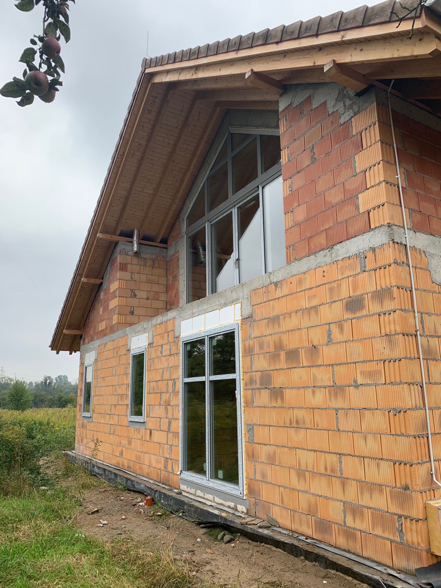 Niedokończony budynek z pustaków ceramicznych z drewnianą konstrukcją dachu i wstawionymi oknami, widoczne detale połączeń murarskich i obróbek okiennych, na pierwszym planie fragment terenu budowy...