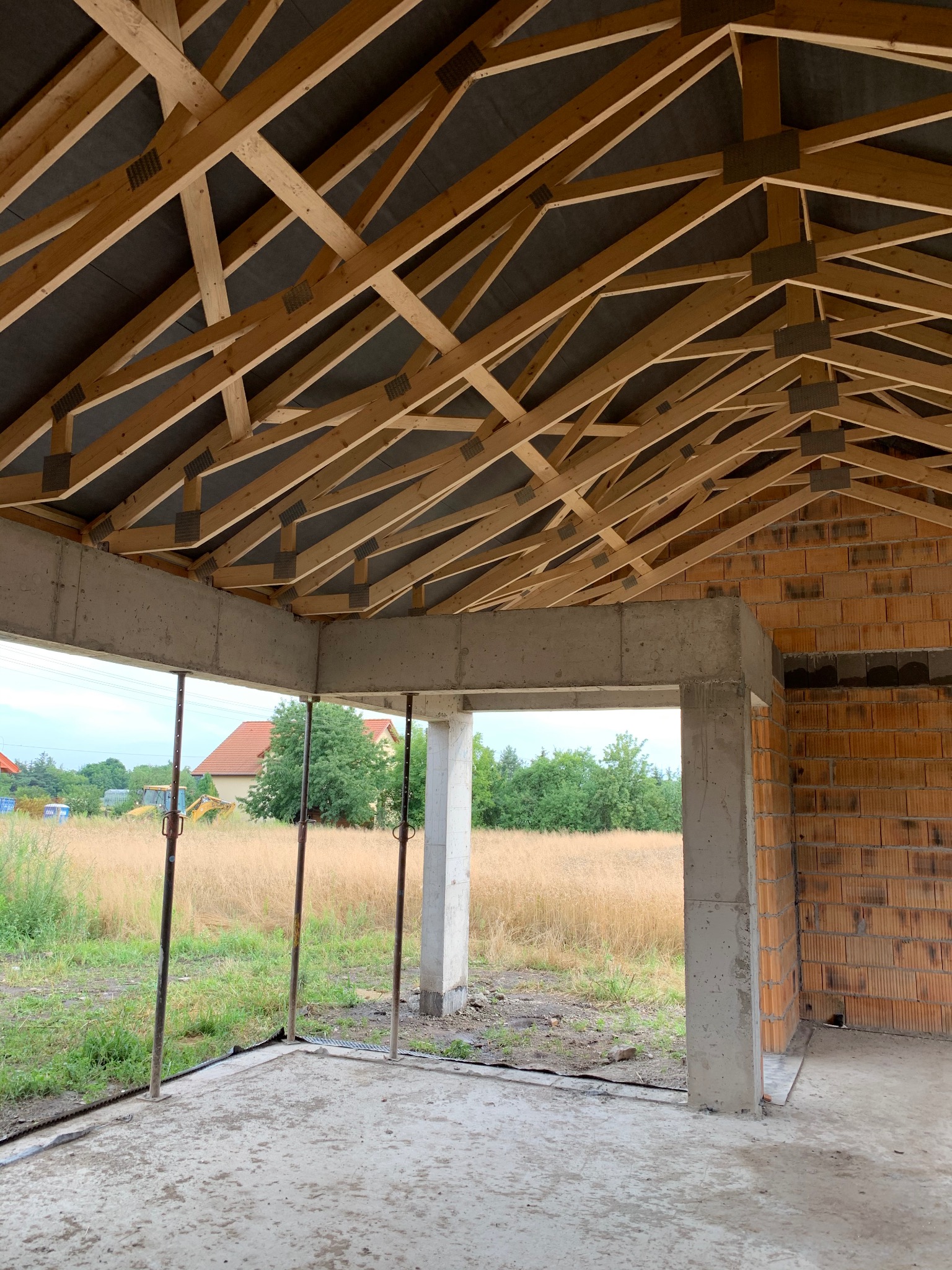 Wnętrze surowego stanu domu w trakcie budowy: widok na drewnianą konstrukcję dachu z czarną membraną, ściany z czerwonej cegły, betonowe słupy i widok na łąkę z żółtą koparką w tle.