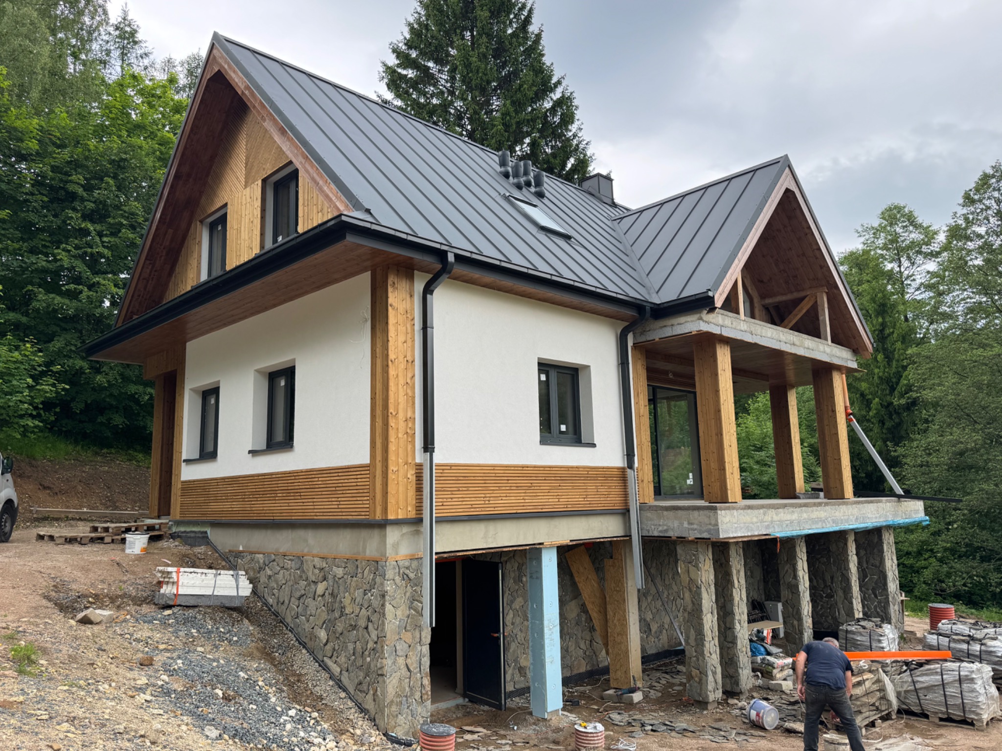 Dwukondygnacyjny dom z dachem z blachy na kamiennej podmurówce, otoczony zielenią. Widoczne elementy drewniane wykończenia i prace wykończeniowe na zewnątrz.