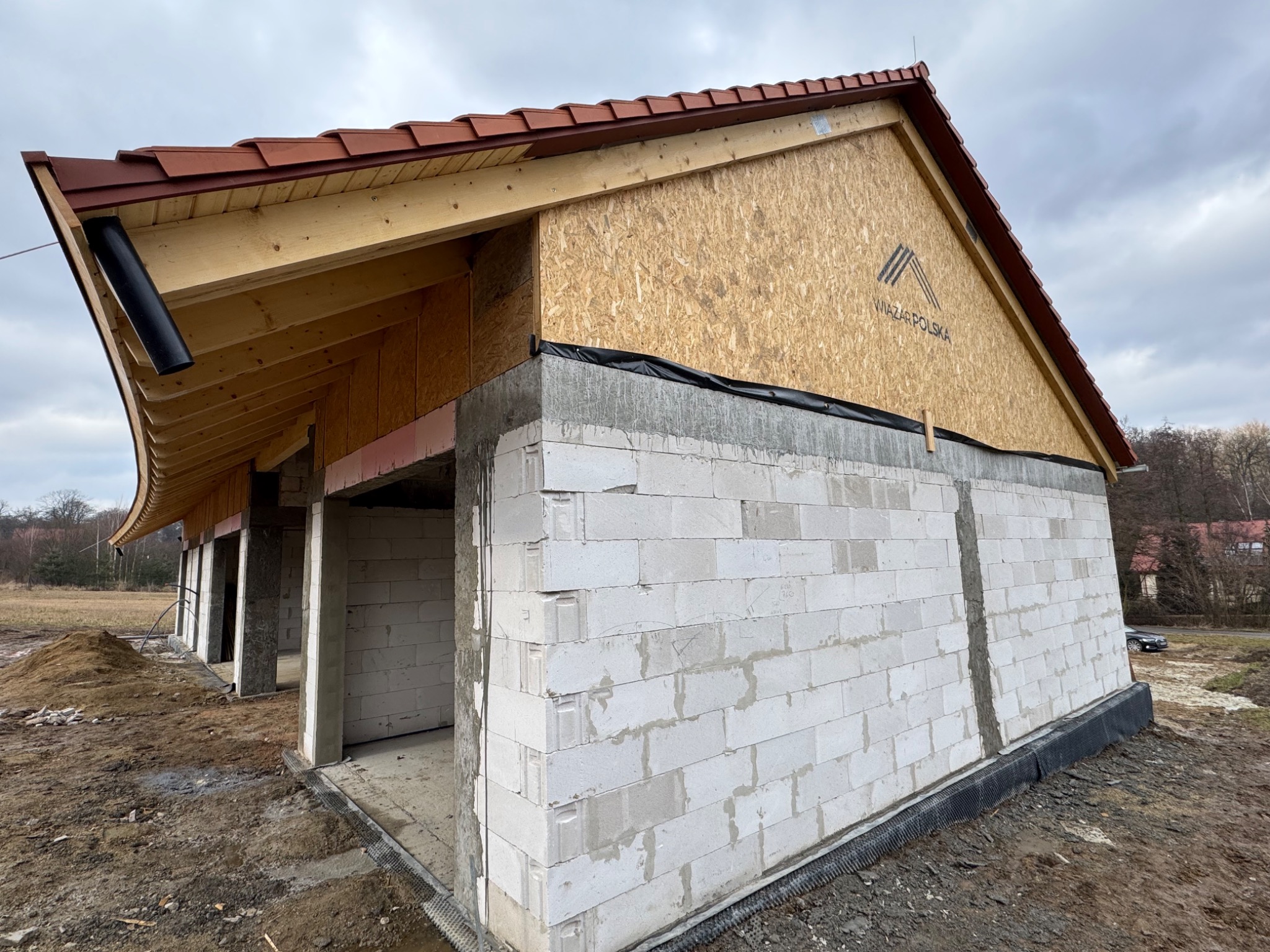 Budowa domu w stanie surowym otwartym: białe bloczki, drewniana konstrukcja dachu z brązową dachówką, widoczne elementy orynnowania. Na ścianie logo producenta.