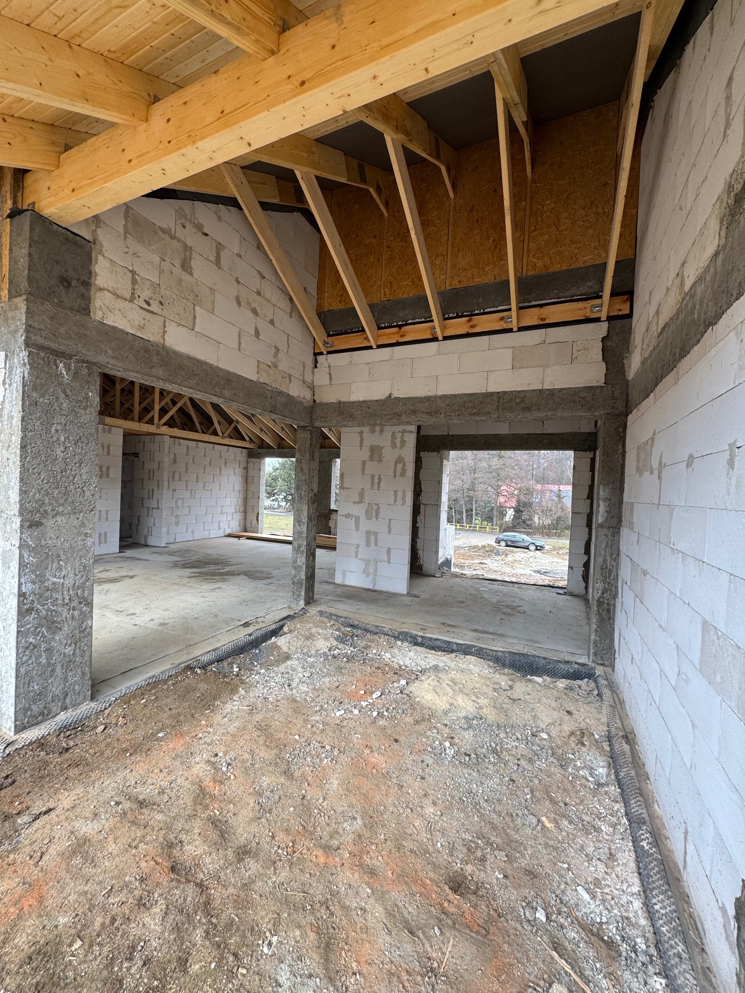 Wnętrze surowego domu z betonu komórkowego w trakcie budowy. Widoczne ściany, betonowe filary, drewniana konstrukcja dachu i ziemia na podłodze. W tle widok na zewnątrz.