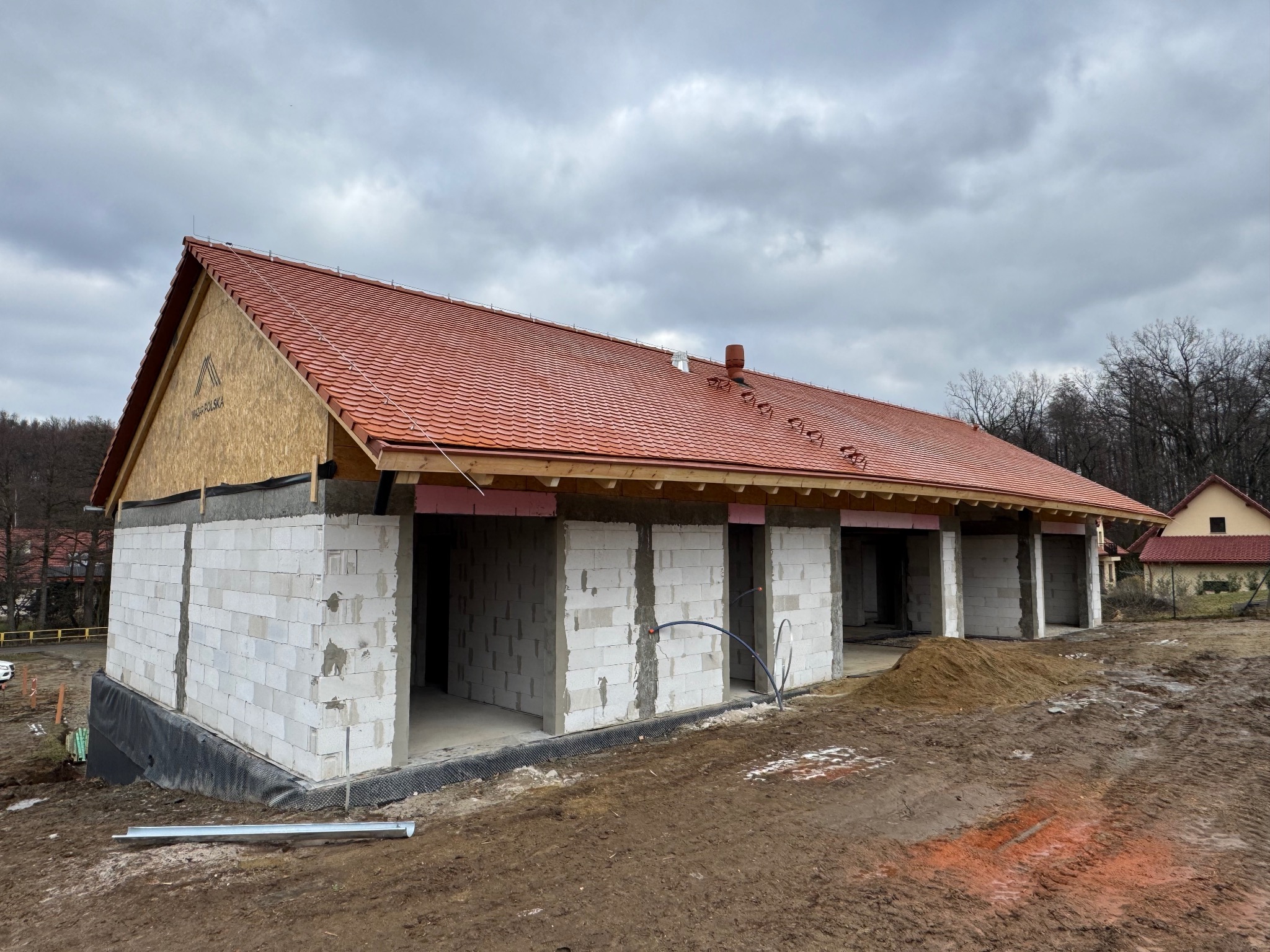 Budowa szeregowca z białych bloczków, z czerwonym dachem w trakcie realizacji. Widoczne elementy konstrukcyjne i przygotowanie terenu. Pochmurne niebo w tle.