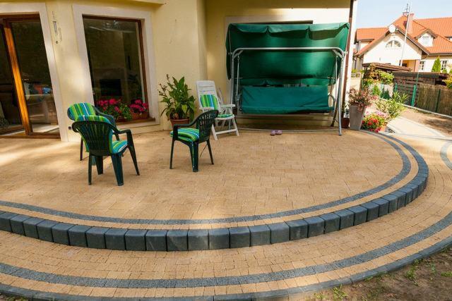 Ułożony taras z kostki brukowej w odcieniach beżu i szarości, z niskim stopniem obrzeżonym ciemnymi kostkami, widoczne meble ogrodowe i huśtawka w tle.