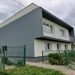 Dwukondygnacyjny dom z szarą elewacją i białymi elementami, balkon z ozdobną balustradą, zielone ogrodzenie i bujna roślinność.