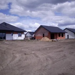 Wczesny etap budowy kilku domów jednorodzinnych: jeden z ocieploną elewacją, drugi w surowej cegle, trzeci z pomalowaną elewacją, na pierwszym planie teren budowy z widocznymi śladami opon