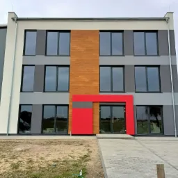 Nowoczesny budynek biurowy z szarą elewacją, drewnianymi panelami i jaskrawoczerwonym obramowaniem wejścia, widok z poziomu gruntu na tle pochmurnego nieba.