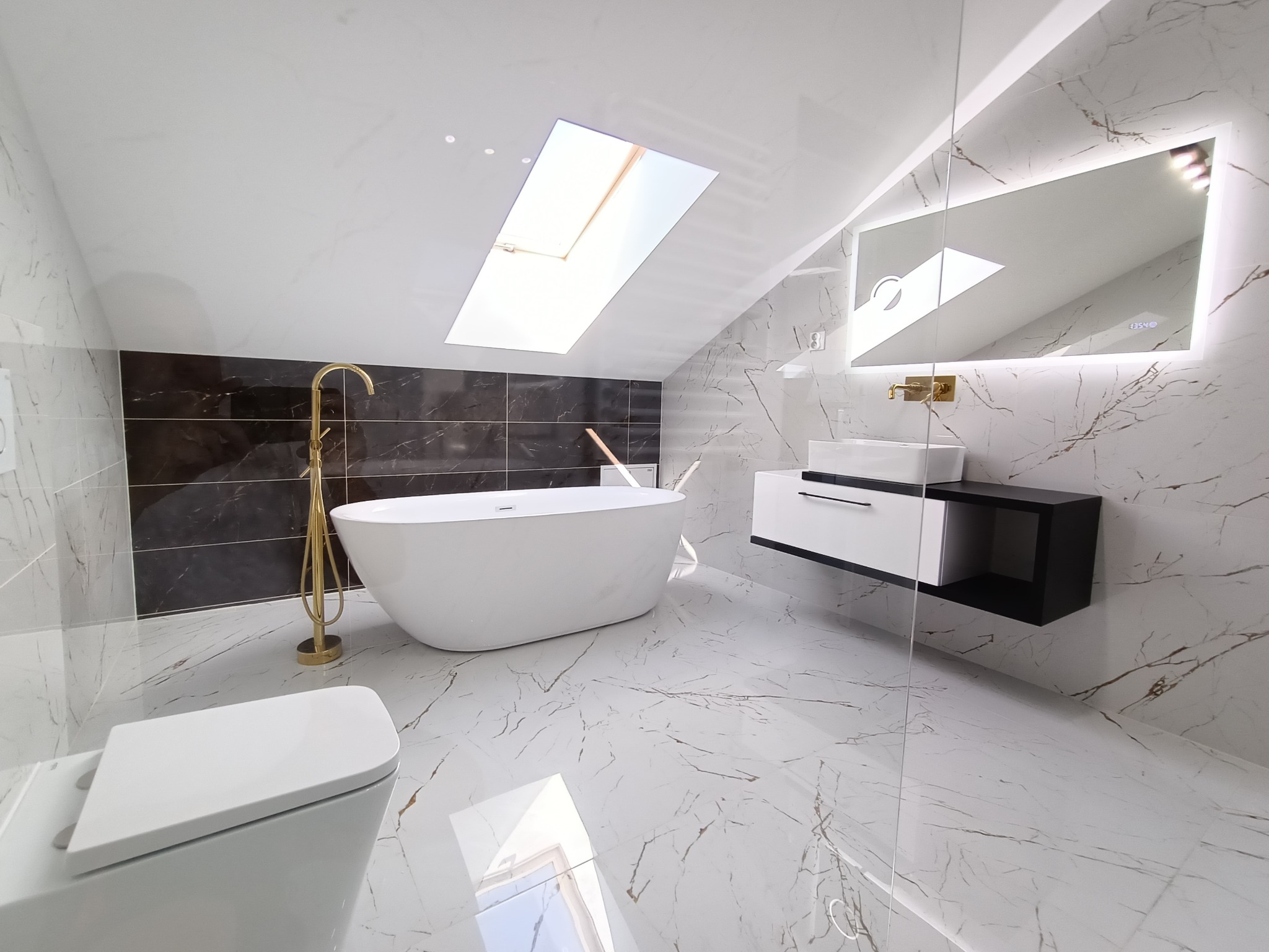 Nowoczesna łazienka na poddaszu z białą wanną wolnostojącą, złotą armaturą, marmurowymi płytkami i oknem dachowym.
