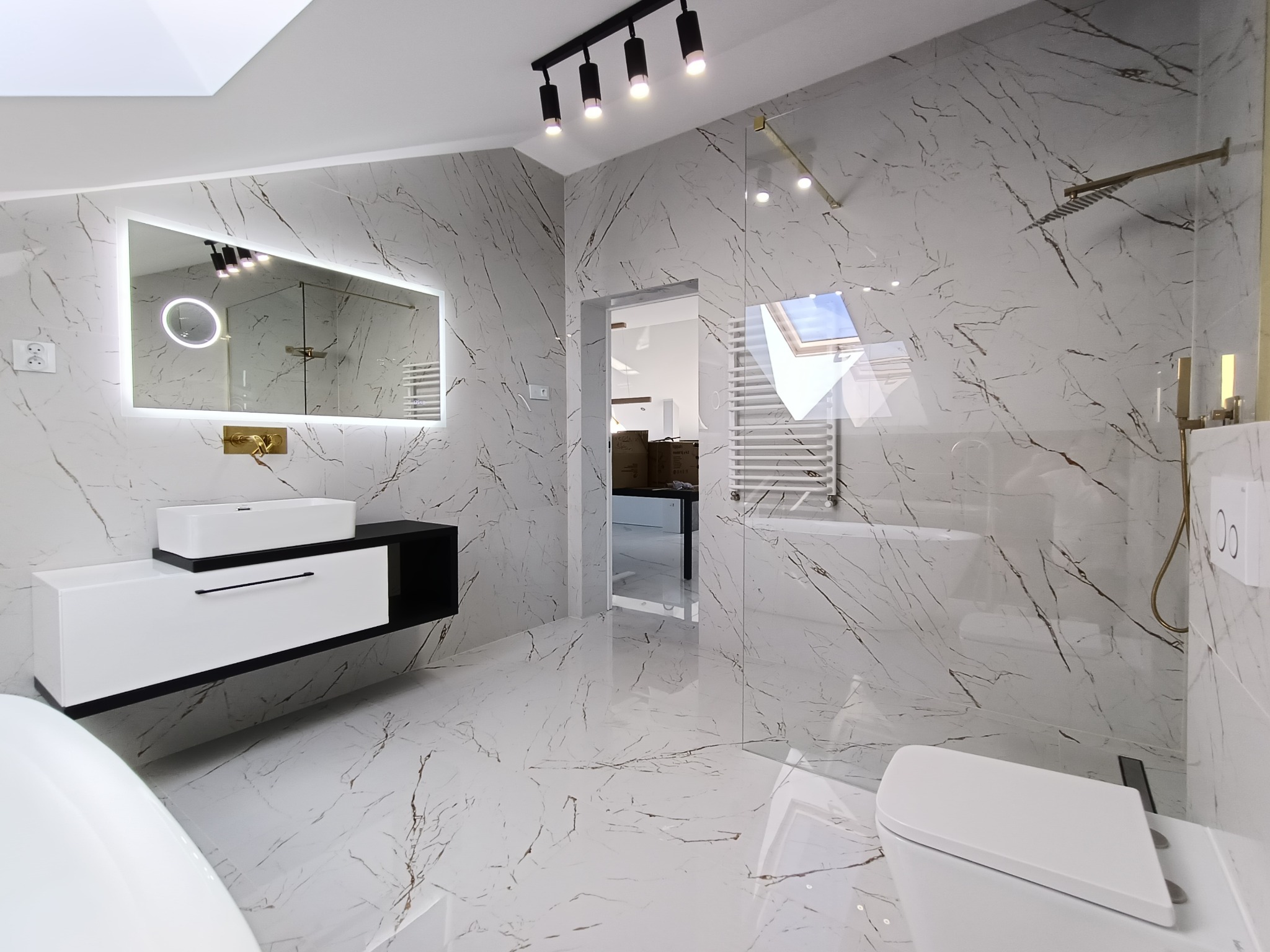 Nowoczesna łazienka z białymi płytkami imitującymi marmur, podświetlanym lustrem, białą szafką z umywalką i szklaną kabiną prysznicową typu walk-in.