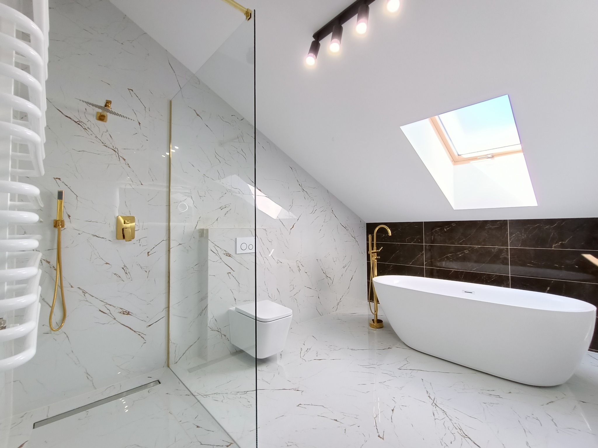 Elegancka łazienka na poddaszu z biało-złotymi marmurowymi płytkami, kabiną prysznicową ze złotymi okuciami, wiszącą toaletą, grzejnikiem drabinkowym i wolnostojącą wanną ze złotą baterią.