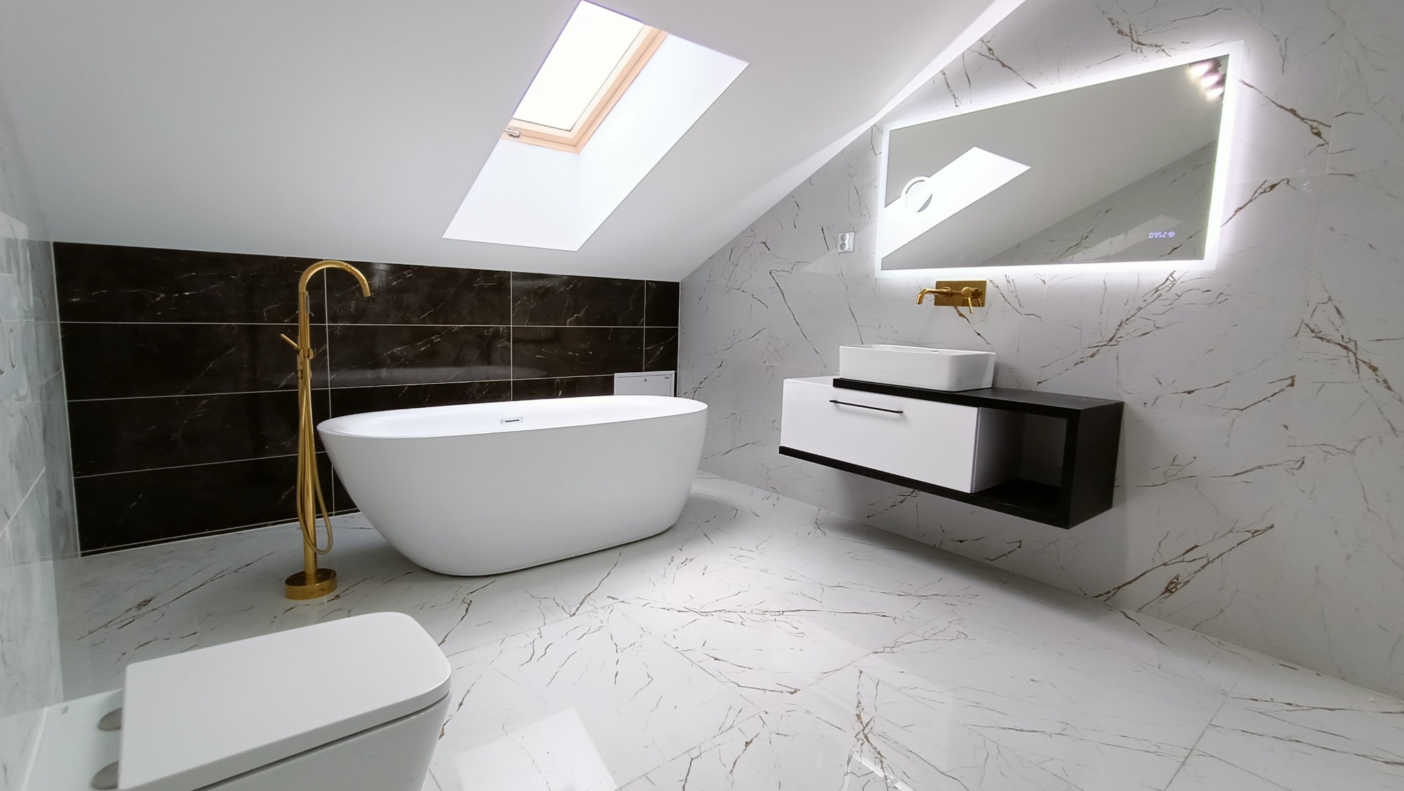 Nowoczesna łazienka na poddaszu z białą wanną wolnostojącą, złotą armaturą, umywalką nablatową na czarno-białej szafce i podświetlanym lustrem na ścianie z marmurowym wzorem.