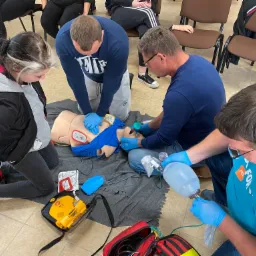 Grupa osób ćwiczy resuscytację krążeniowo-oddechową na fantomie w sali szkoleniowej. Widoczny defibrylator, maska twarzowa i worek ambu.