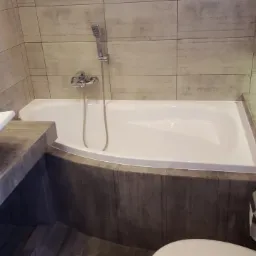 Nowoczesna łazienka z wanną, umywalką i toaletą w szarościach. Charakterystyczne płytki imitujące drewno na ścianach i obudowie wanny. Bateria umywalkowa z chromu.