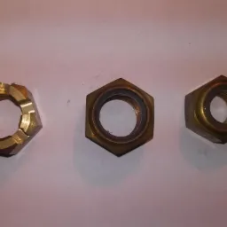 Trzy mosiężne nakrętki: z lewej koronowa, po środku standardowa sześciokątna z pierścieniem, z prawej sześciokątna z wkładką polimerową.