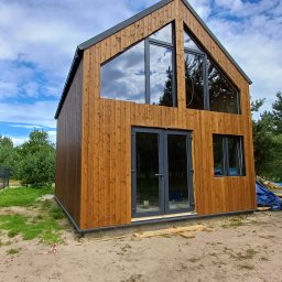 Woodenfactory Sp. z o.o. - Nowoczesny, dwukondygnacyjny dom szkieletowy z drewnianą elewacją, dużymi oknami i szarą stolarką, na tle błękitnego nieba z chmurami.