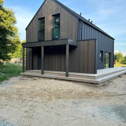 Woodenfactory Sp. z o.o. - Nowoczesny, ciemny dom z drewna z podestem wejściowym. Widoczne pionowe deski, duże okna i zadaszone wejście. Otoczenie w stanie surowym.