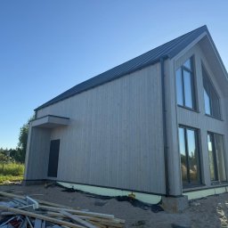 Woodenfactory Sp. z o.o. - Nowoczesny dom szkieletowy z drewnianą elewacją i czarnym dachem, widoczny z zewnątrz w słoneczny dzień, z elementami placu budowy u podstawy.