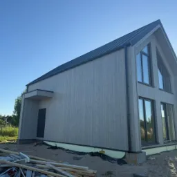 Nowoczesny dom szkieletowy z drewnianą elewacją i czarnym dachem, widoczny z zewnątrz w słoneczny dzień, z elementami placu budowy u podstawy.