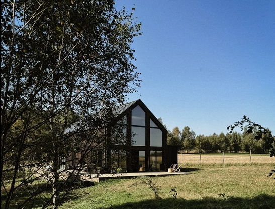 Czarna, nowoczesna stodoła z dużymi oknami na tle łąki i lasu, widziana przez gałęzie drzewa. Minimalistyczny design, jasne niebo.