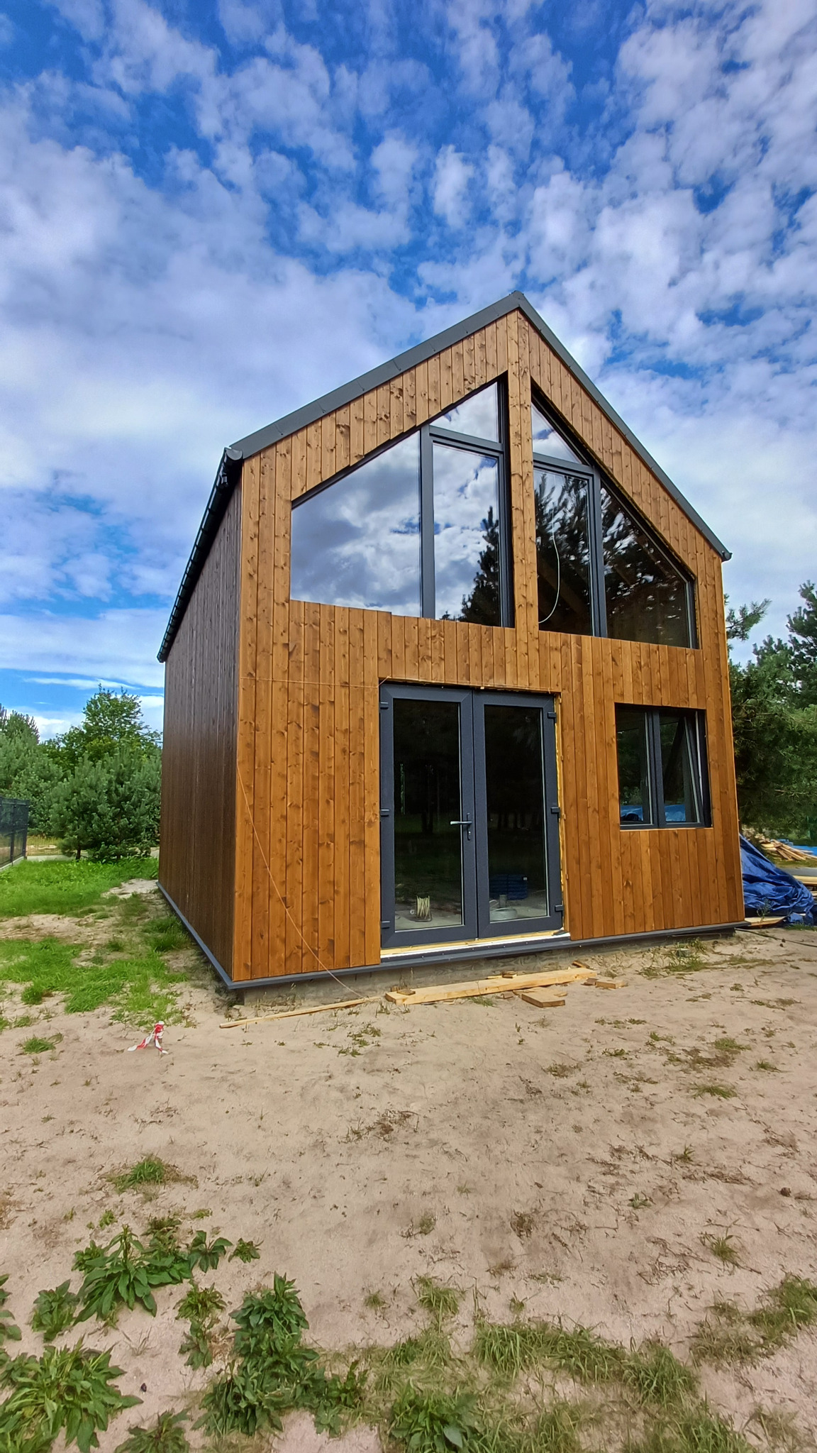 Nowoczesny, dwukondygnacyjny dom szkieletowy z drewnianą elewacją, dużymi oknami i szarą stolarką, na tle błękitnego nieba z chmurami.
