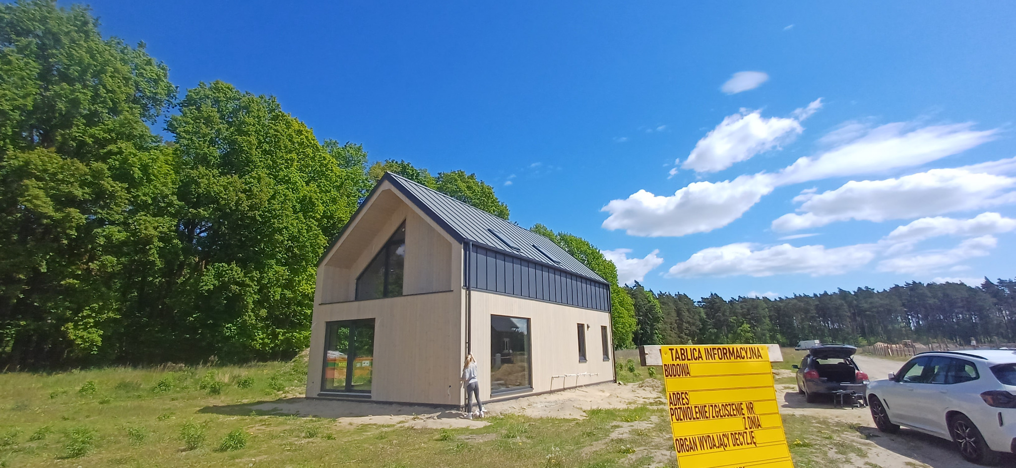 Nowoczesny dom z jasnego drewna z ciemnym dachem na tle lasu i błękitnego nieba. Kobieta stoi przy ścianie, obok tablica informacyjna budowy i zaparkowane auto.