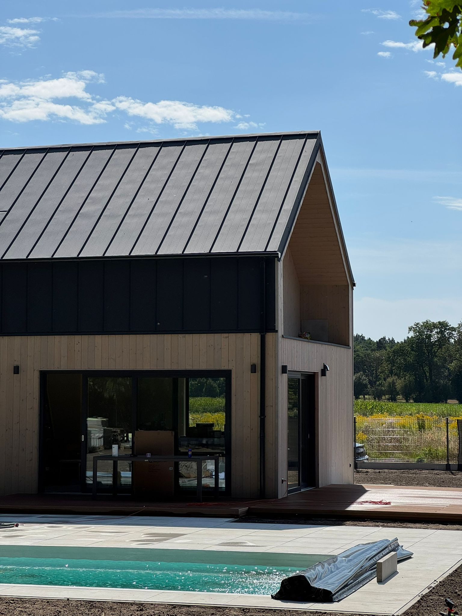 Nowoczesny dom z jasną elewacją, ciemnym dachem i basenem w ogrodzie. Widoczny taras z drewna i duże przesuwne okna. Budynek w trakcie wykańczania.