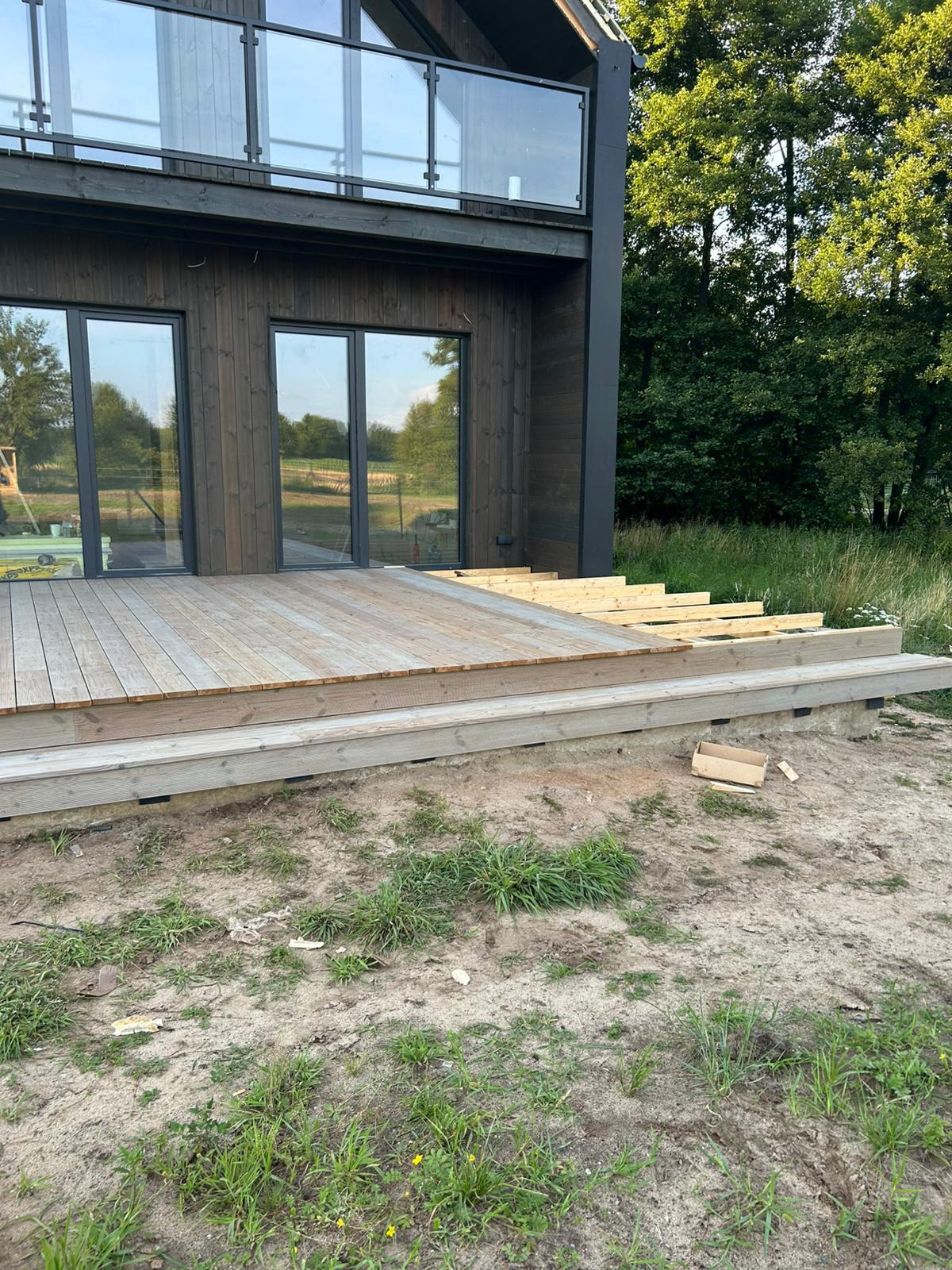 Drewniany taras w trakcie budowy przy nowoczesnym domu z ciemnego drewna i szklaną balustradą. Widoczne deski, podkonstrukcja i otoczenie z zielenią.