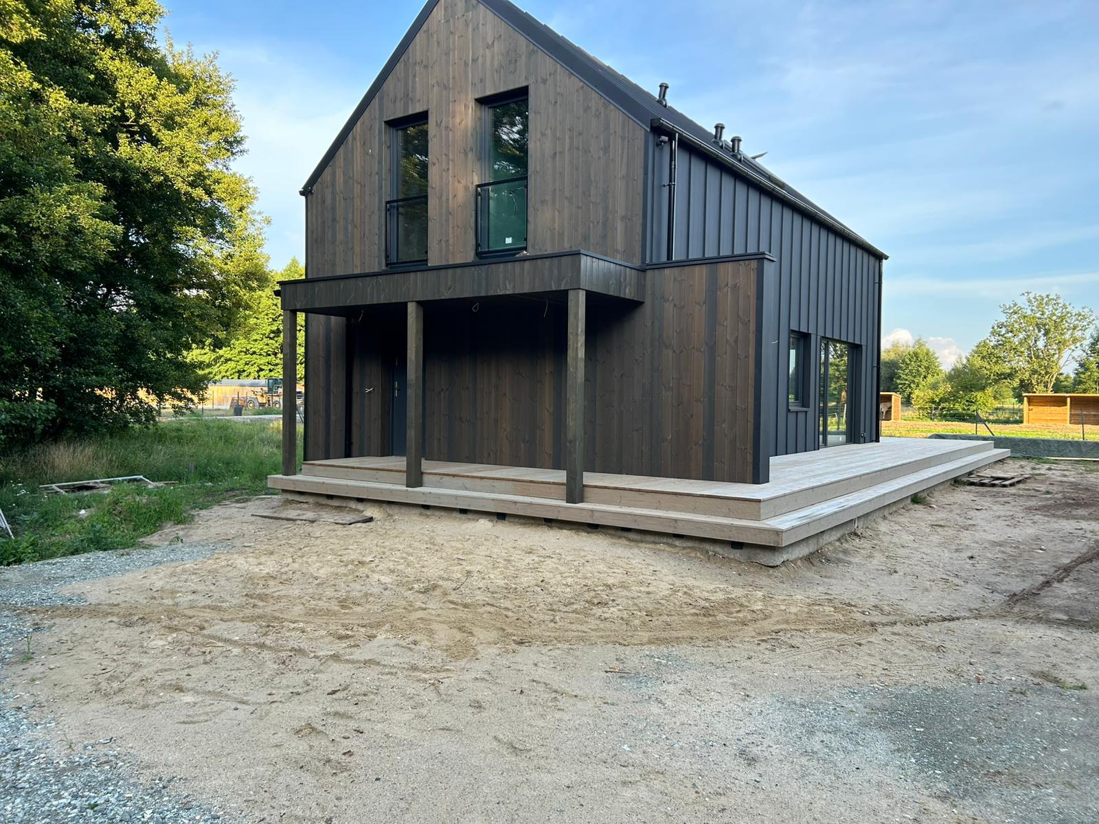 Nowoczesny, ciemny dom z drewna z podestem wejściowym. Widoczne pionowe deski, duże okna i zadaszone wejście. Otoczenie w stanie surowym.