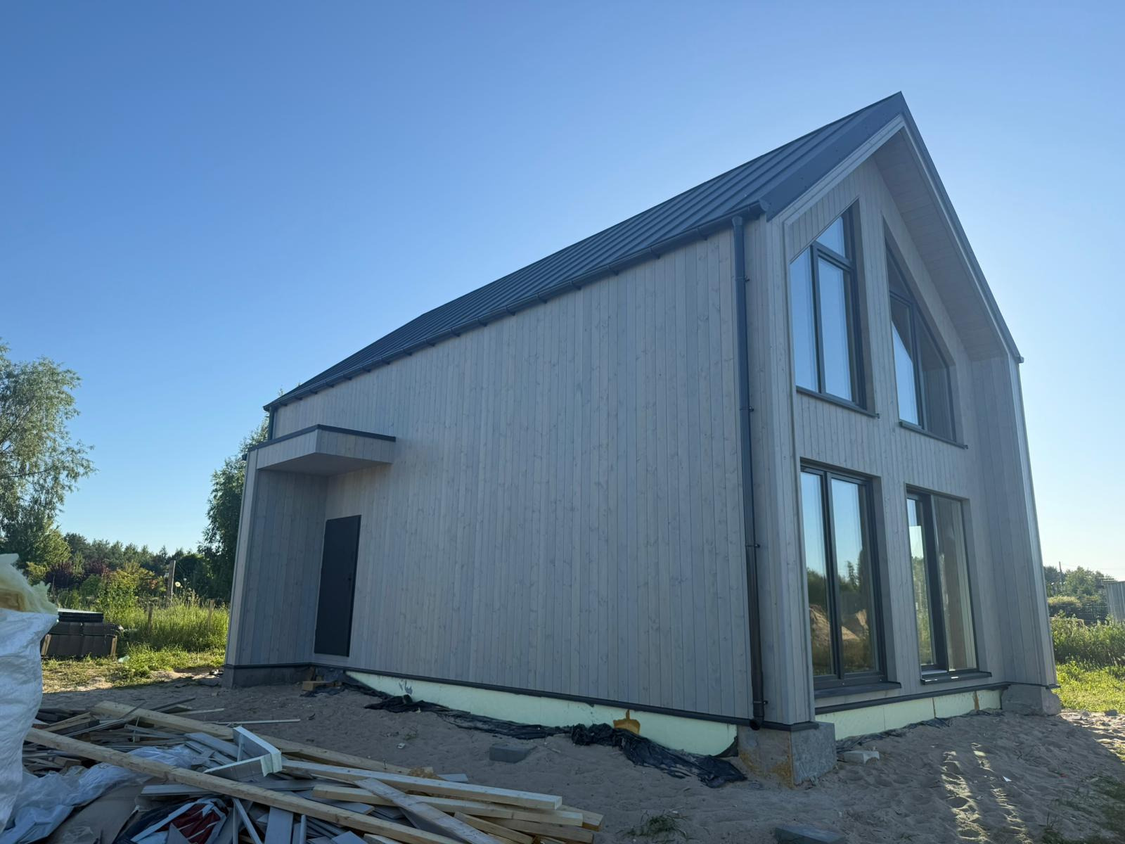 Nowoczesny dom szkieletowy z drewnianą elewacją i czarnym dachem, widoczny z zewnątrz w słoneczny dzień, z elementami placu budowy u podstawy.
