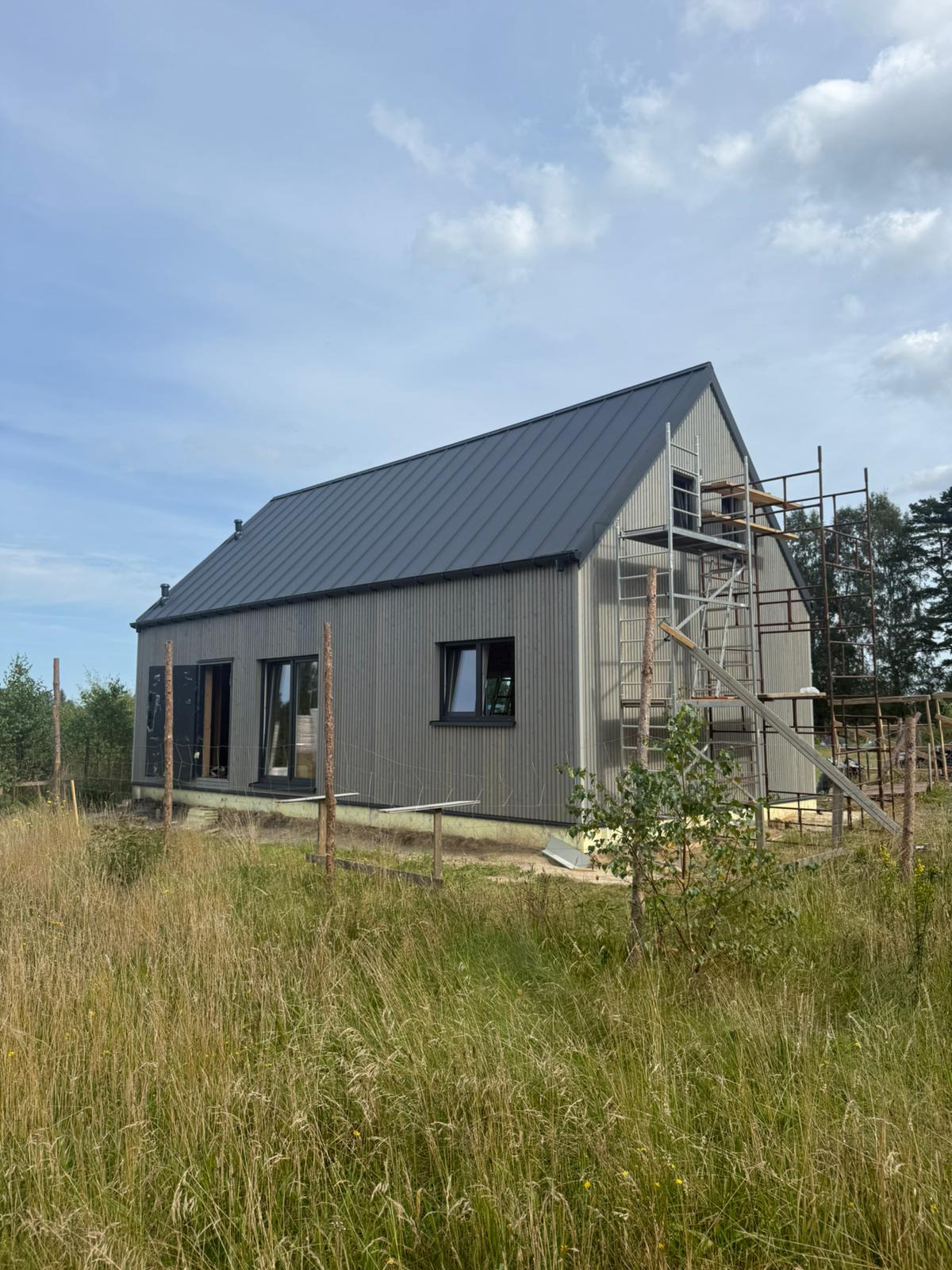 Szary dom z blachy na etapie budowy, widoczne rusztowania i wysoka trawa. Uwagę przykuwa prosta bryła budynku i duże okna z czarnymi ramami.