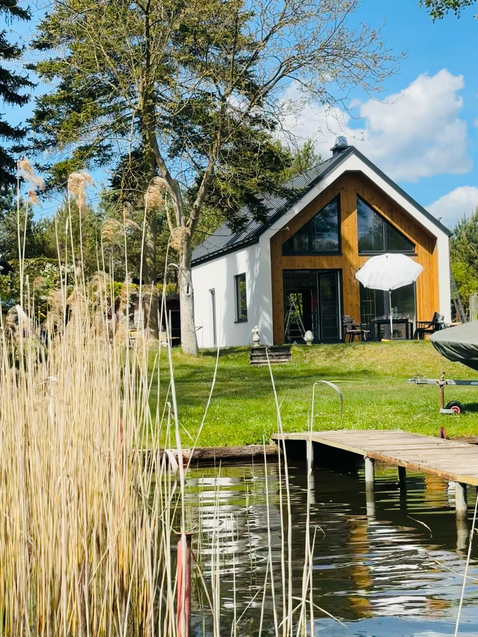 Nowoczesny dom z drewnianą fasadą i dużymi oknami, widoczny z perspektywy pomostu nad jeziorem, otoczony zielenią i trzcinami. Biała elewacja.