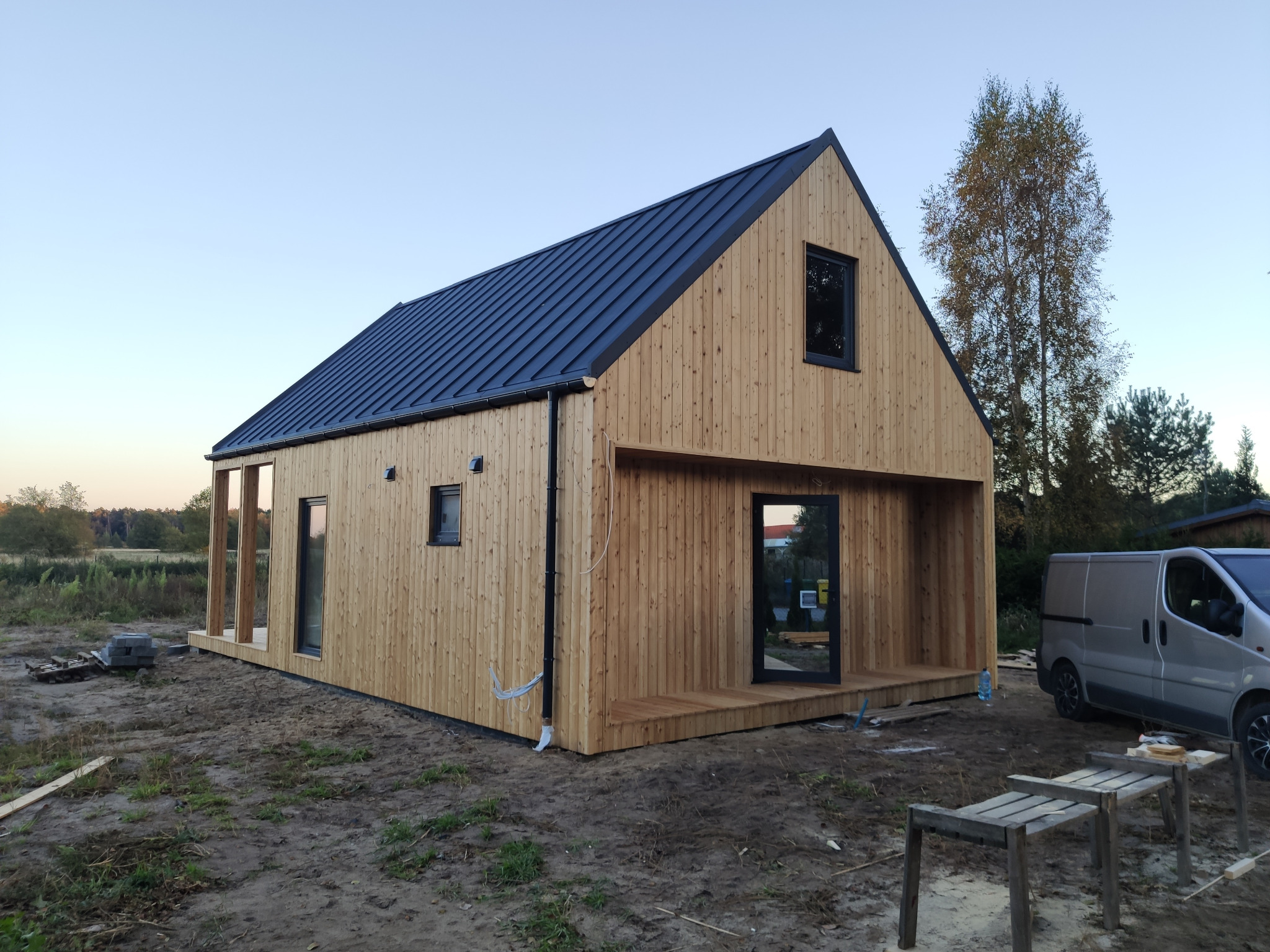 Surowy, drewniany dom z ciemnym, metalowym dachem i dużymi oknami, częściowo otwartą przestrzenią i zaparkowanym obok samochodem dostawczym na działce w trakcie zagospodarowywania.