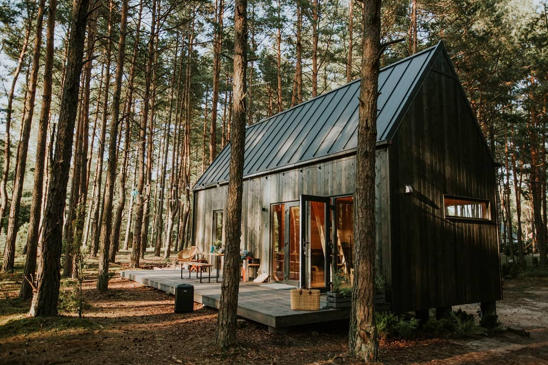 Czarny, drewniany domek letniskowy z dwuspadowym dachem, otoczony wysokimi sosnami, z widocznym tarasem i otwartymi, przeszklonymi drzwiami balkonowymi.