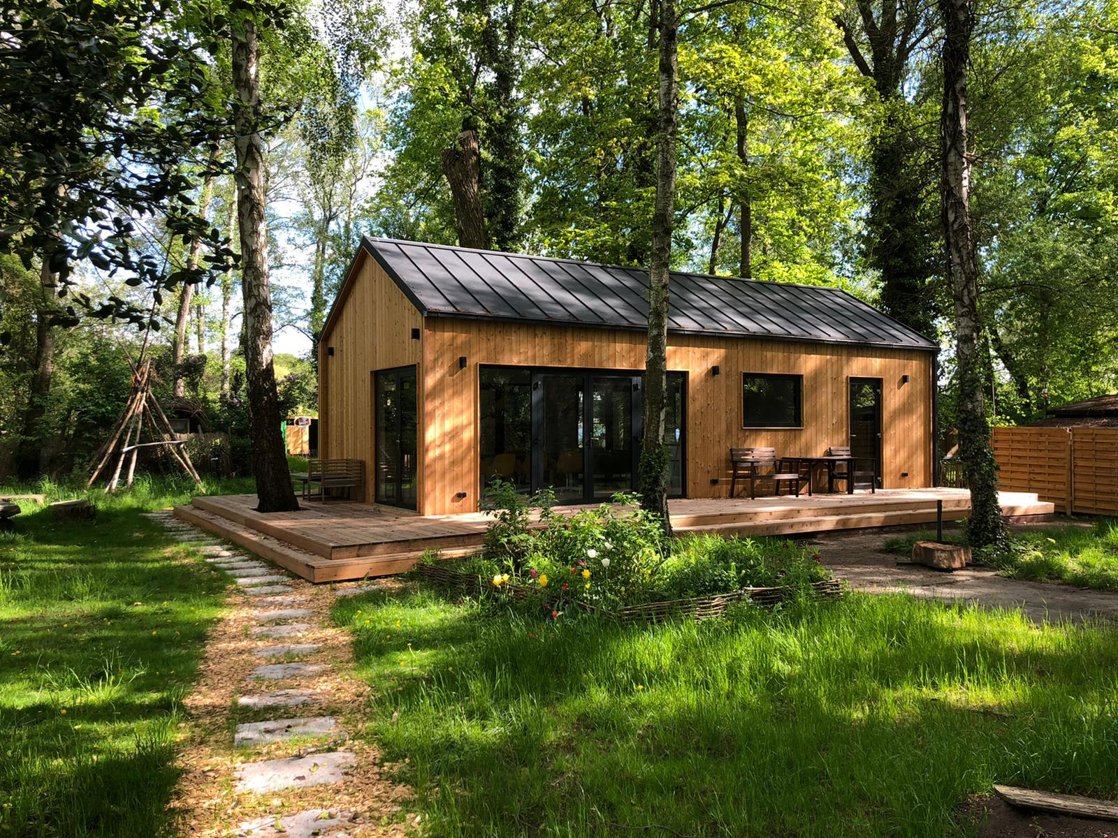 Nowoczesny, drewniany dom z czarnym dachem, otoczony zielenią i drzewami, z tarasem i ścieżką z kamieni.