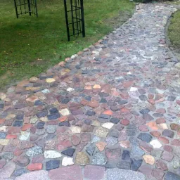 Ścieżka z nieregularnej kostki granitowej w ogrodzie, z widocznymi elementami metalowej konstrukcji w tle i mokrą nawierzchnią po deszczu. Układanka w odcieniach szarości, czerwieni i brązu.