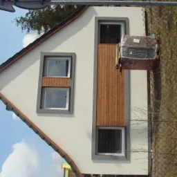 Dwukondygnacyjny dom z jasną elewacją, szarą stolarką okienną i drewnianymi elementami dekoracyjnymi, widok z zewnątrz, obok domu składowane materiały budowlane.