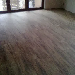 Ułożona podłoga z płytek imitujących drewno, widok z niskiego kąta, z oknem balkonowym w tle i surową ścianą.