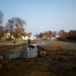 Teren budowy z przygotowanymi betonowymi blokami fundamentowymi na palecie i stosem metalowych prętów, widoczny mężczyzna w oddali oraz drzewa w jesiennej scenerii.