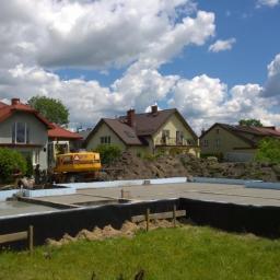 KAMIL WILCZYŃSKI ARCHITEKTURA I BUDOWNICTWO - Wylany fundament pod budowę domu jednorodzinnego, zabezpieczony czarną folią i styropianem, z widoczną żółtą koparką i stertą ziemi w tle oraz zabudową mieszkalną.
