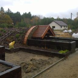 KAMIL WILCZYŃSKI ARCHITEKTURA I BUDOWNICTWO - Krajobraz z ziemianką częściowo wkopaną w ziemię, z drewnianym zadaszeniem i schodami, otoczoną podwyższonymi grządkami i workami ziemi, w tle dom i las.