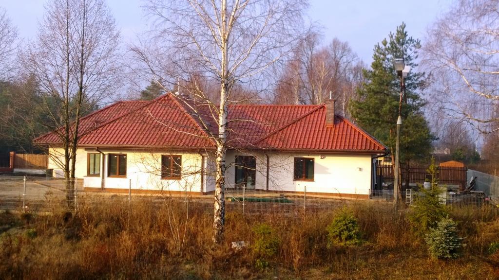 Parterowy dom jednorodzinny z czerwoną dachówką, białą elewacją i ciemnymi oknami, otoczony siatką ogrodzeniową i drzewami bez liści, sfotografowany wczesną wiosną.