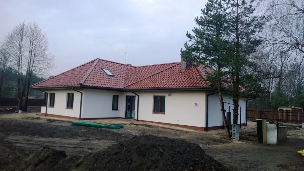 Parterowy budynek z czerwonym dachem, otynkowany na biało, z widocznymi brązowymi oknami i drzwiami, na działce w trakcie zagospodarowywania.