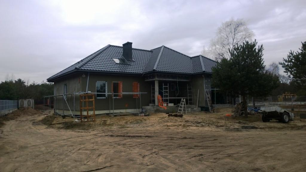 Budowa domu jednorodzinnego w stanie surowym zamkniętym, z ciemną dachówką, widoczne rusztowania i teren budowy.
