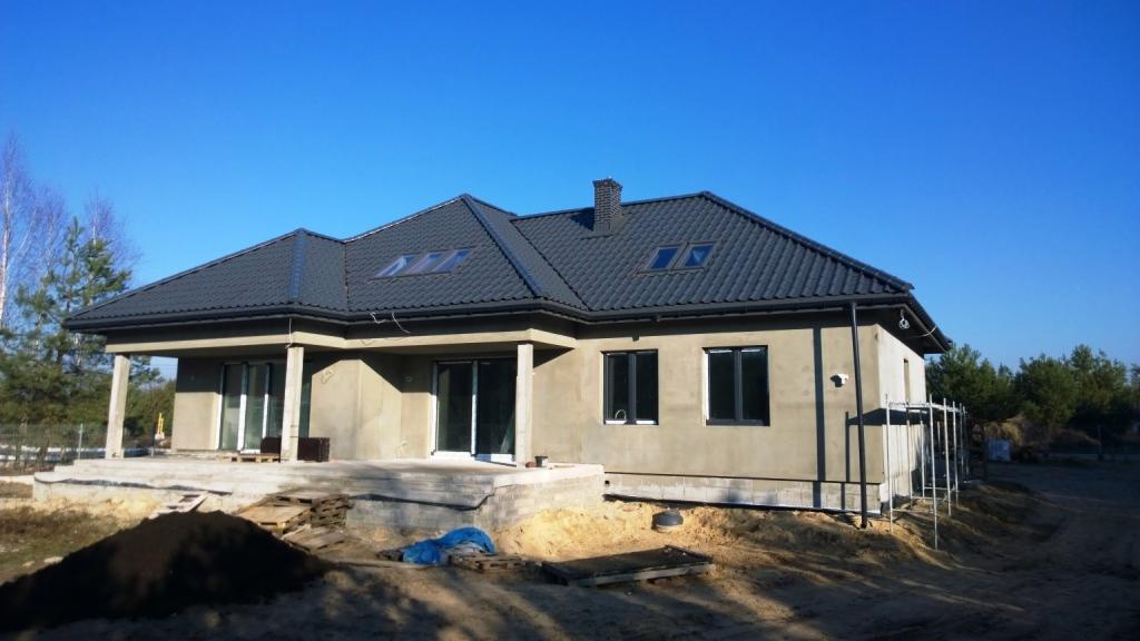 Surowy, parterowy dom murowany z ciemną dachówką i lukarnami w trakcie budowy, widoczne rusztowania i niezagospodarowany teren.