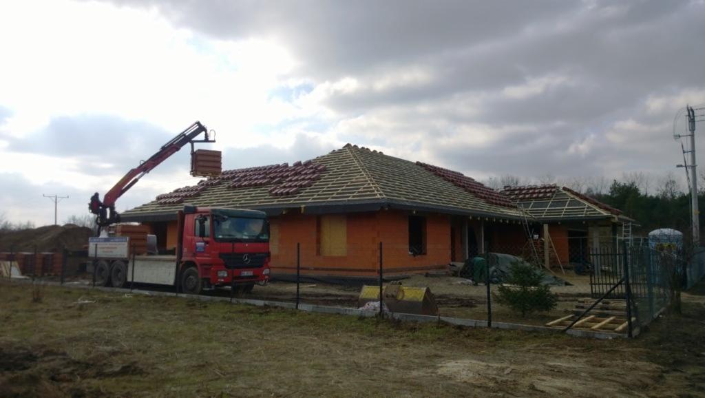 Czerwony dźwig samochodowy dostarcza paletę z czerwoną dachówką na drewnianą konstrukcję dachu powstającego parterowego domu z pomarańczowej cegły.