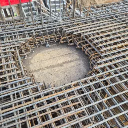 Siatka z prętów zbrojeniowych ułożona na deskowaniu, przygotowana do zalania betonem, widok z góry, fragment czerwonej barierki ochronnej w tle.