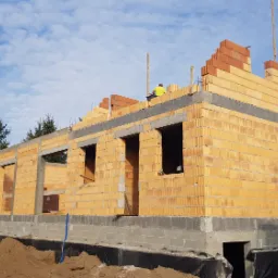 Wznoszenie ścian zewnętrznych z pustaków ceramicznych Porotherm na fundamencie z betonu komórkowego, widoczny robotnik w żółtej kamizelce na koronie muru oraz minikoparka Atlas Weycor na pierwszym...